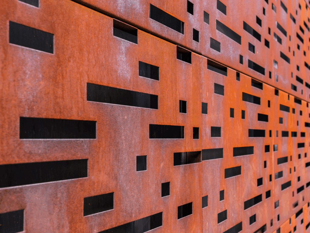 Close-up of a rust-colored metal wall with rectangular cutouts of various sizes, creating a geometric pattern with black spaces visible behind the panels.