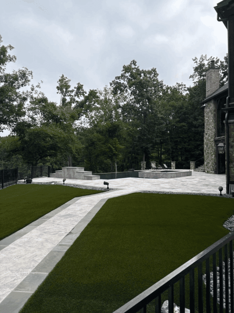 A paved stone walkway divides two sections of artificial grass in a backyard, with a stone patio and pool area surrounded by trees in the background.