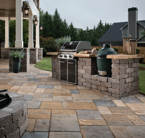 Outdoor patio with stone flooring, a built-in grill, green ceramic smoker, countertop, potted plant, and covered seating area; trees and a house in the background.