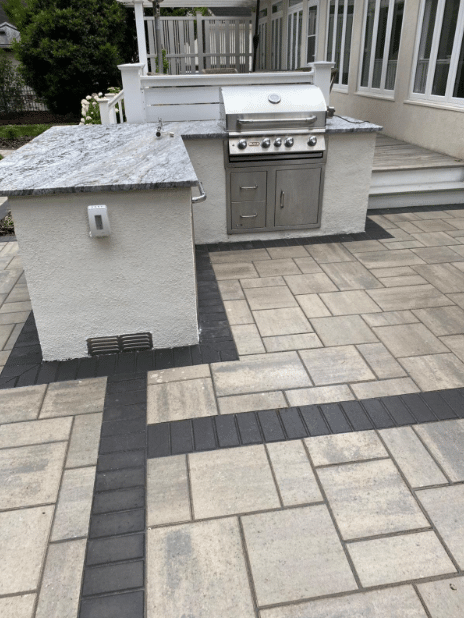 Outdoor kitchen with a stainless steel grill and marble countertop on a patio with light and dark rectangular pavers.