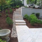 Stone steps lead up a landscaped slope with mulch, shrubs, and a row of rocks; a decorative planter and patio furniture are visible in the foreground.