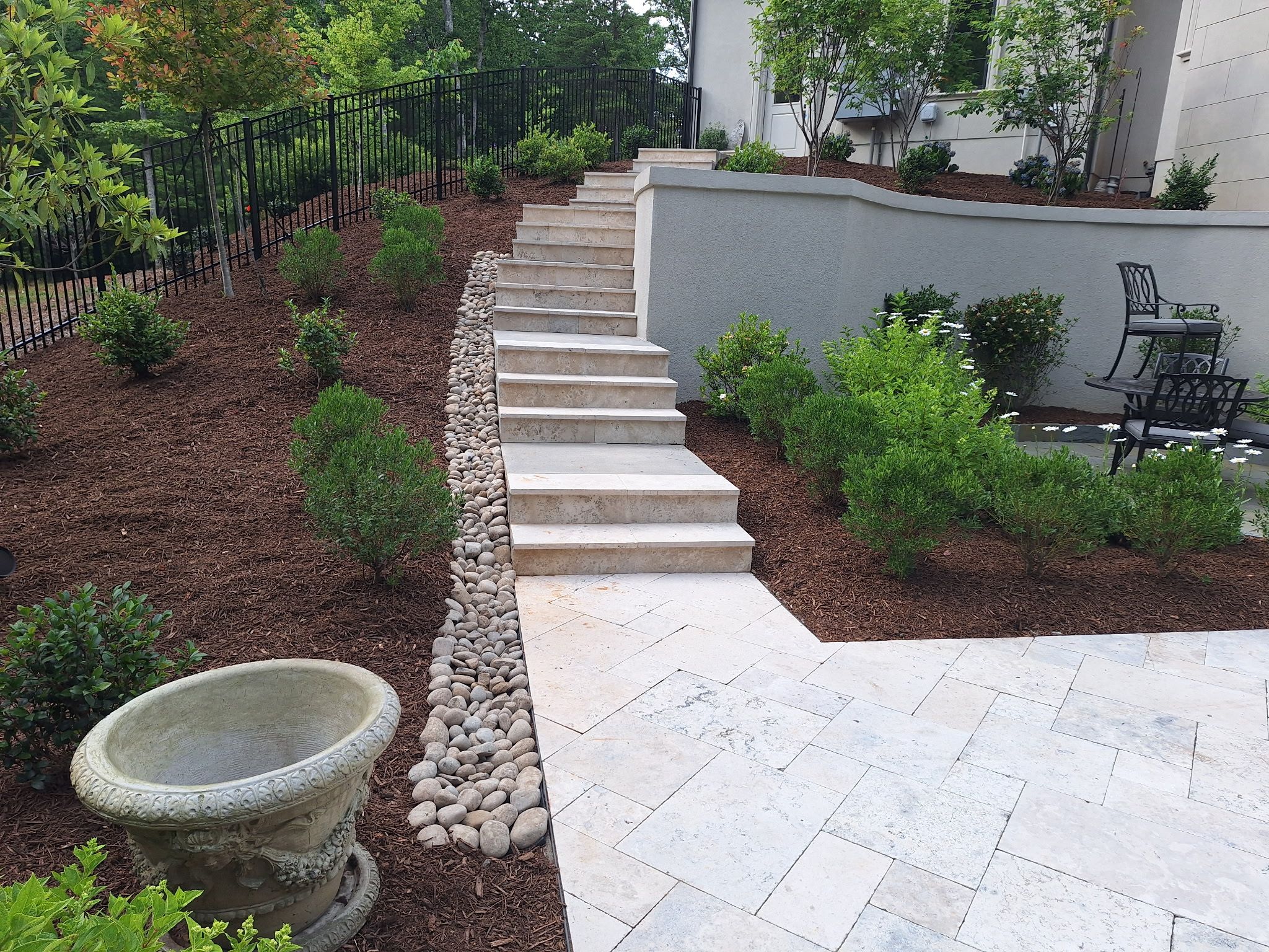 Stone steps lead up a landscaped slope with mulch, shrubs, and a row of rocks; a decorative planter and patio furniture are visible in the foreground.