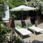 Two white lounge chairs with green and blue pillows sit under a white patio umbrella on a stone patio, surrounded by lush greenery and blooming bushes.