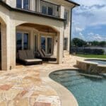 A backyard patio with lounge chairs, a hot tub, and a swimming pool overlooking a lake, bordered by a wrought iron fence under a partly cloudy sky.