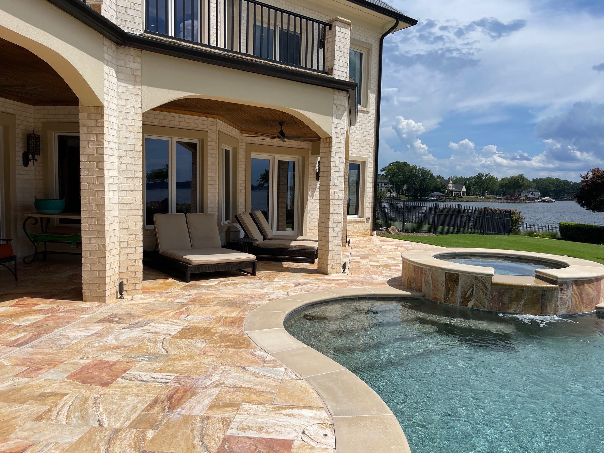 A backyard patio with lounge chairs, a hot tub, and a swimming pool overlooking a lake, bordered by a wrought iron fence under a partly cloudy sky.