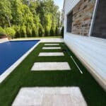 Rectangular stone pavers are set in artificial grass alongside a pool next to a white house with stone accents; a pool net rests by the wall.