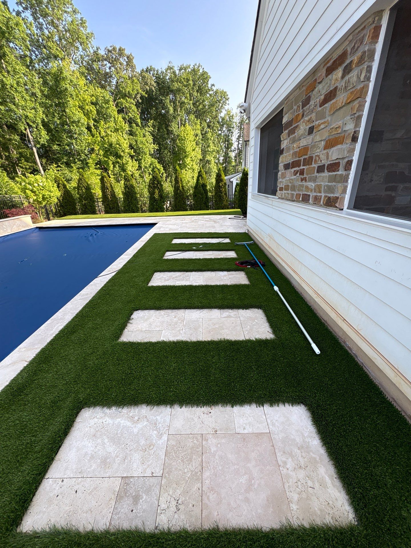Rectangular stone pavers are set in artificial grass alongside a pool next to a white house with stone accents; a pool net rests by the wall.