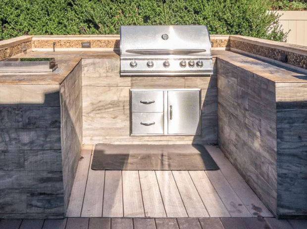 Outdoor built-in stainless steel gas grill with storage drawers and cabinets, set in a stone countertop with surrounding greenery in the background.