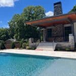A modern house with a stone patio features wide steps leading down to a clear swimming pool, surrounded by trees and neighboring homes under a blue sky.