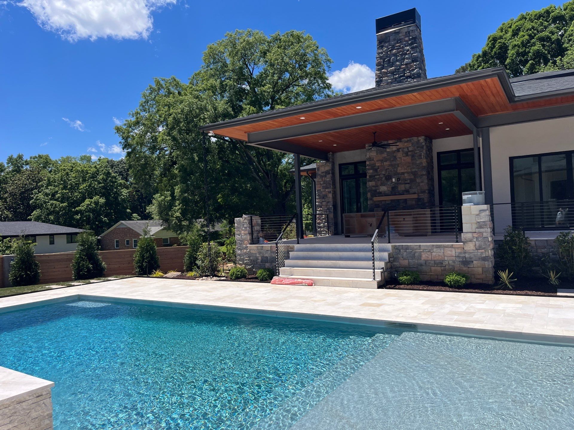 A modern house with a stone patio features wide steps leading down to a clear swimming pool, surrounded by trees and neighboring homes under a blue sky.
