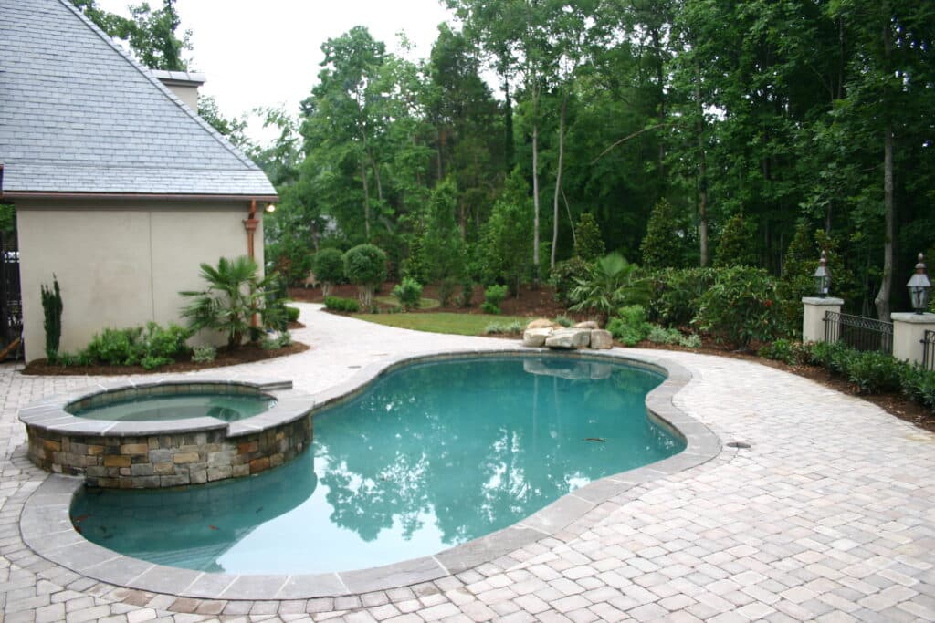 A backyard with a stone-bordered swimming pool and attached hot tub, surrounded by a paved patio and landscaped greenery.