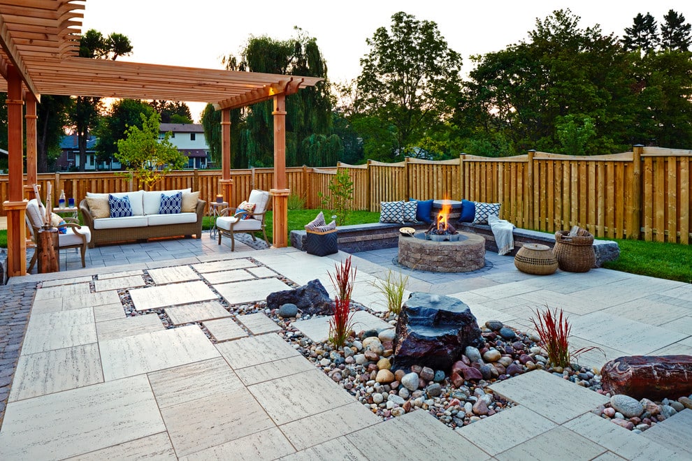 A backyard patio with a wooden pergola, outdoor furniture, a stone fire pit, decorative rocks, and a wooden fence, surrounded by trees and greenery at sunset.