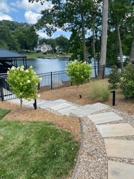 Stone walkway with steps leads down to a lakeside fence, flanked by green shrubs and trees, with a dock and houses visible across the water.