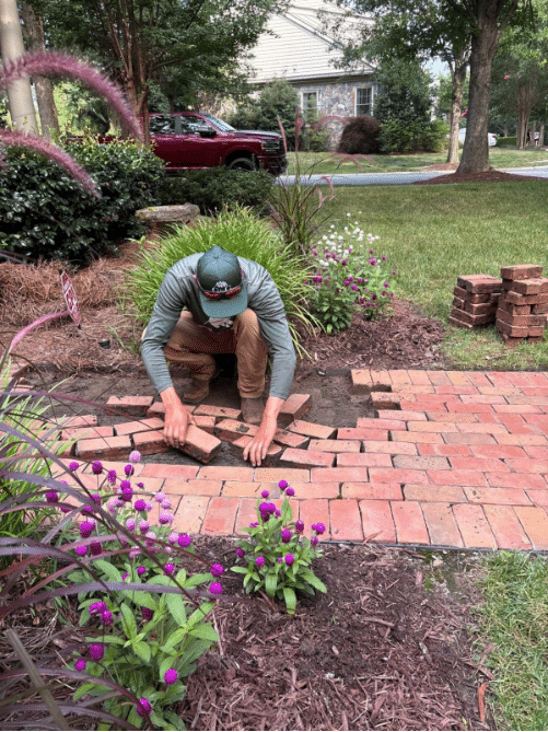 A person kneels on the ground, arranging bricks to repair or build a walkway in a garden with flowers and green plants nearby.