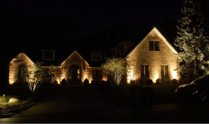 A large brick house is illuminated by exterior lights at night, highlighting the facade and surrounding trees, with a driveway visible in front.