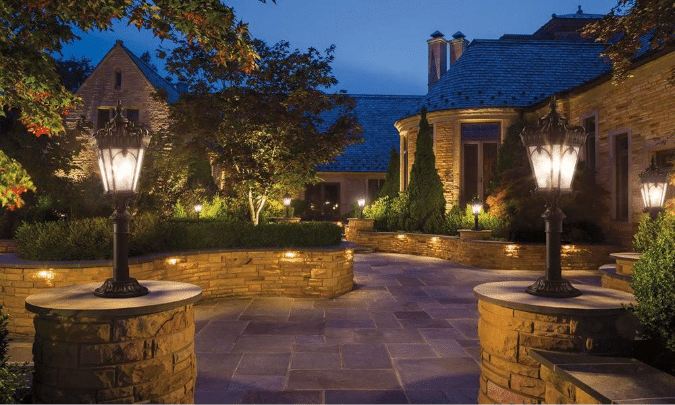 Stone pathway and courtyard of a house at dusk, illuminated by large decorative lamp posts and landscape lighting, surrounded by manicured greenery and stone walls.