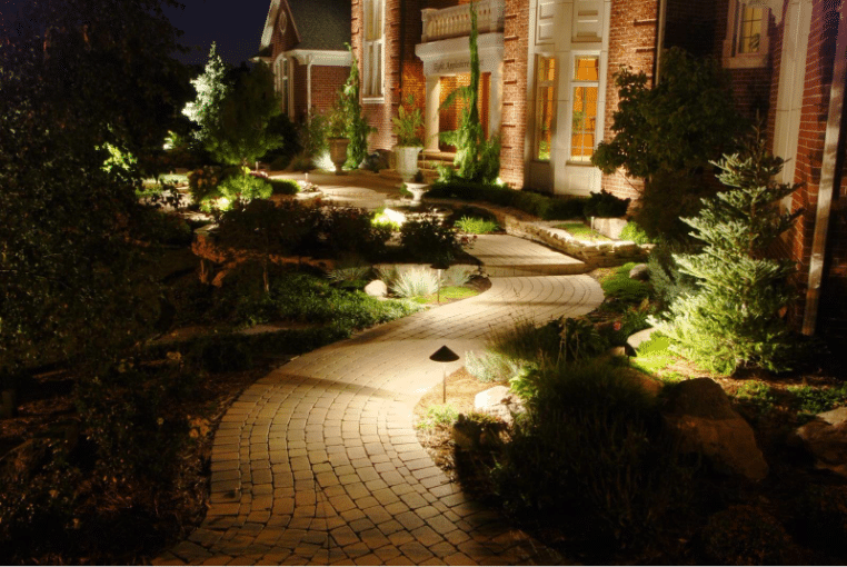 Curved stone pathway with garden lighting leading to a brick house entrance at night, surrounded by shrubs and landscaped plants.