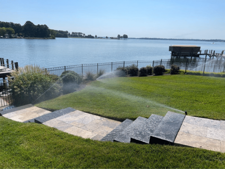 Sprinklers watering a lawn with stone steps, near a fenced waterfront area and a dock on a sunny day.