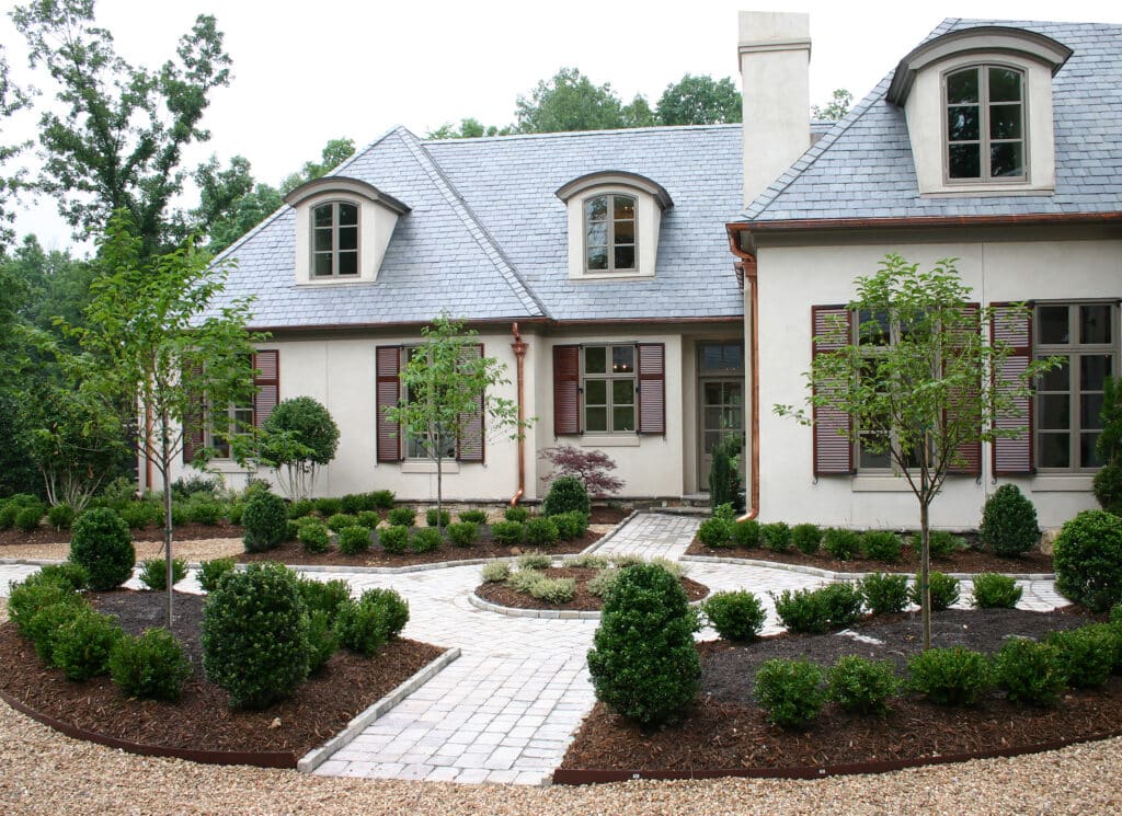 A large house with a gray roof, dormer windows, and red shutters is surrounded by neatly landscaped bushes, trees, and a brick walkway.