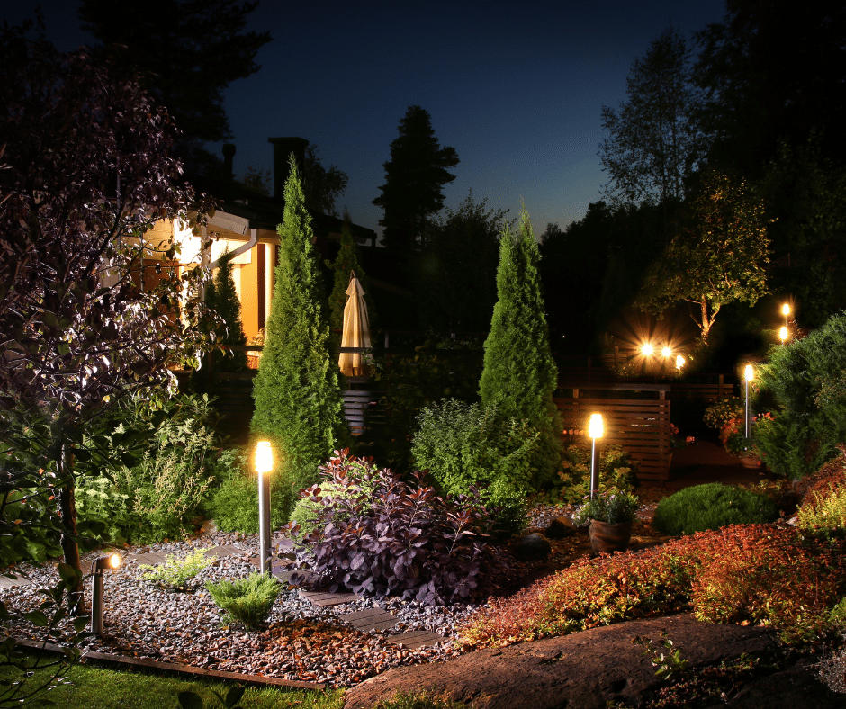 A well-lit garden at night features various trees, shrubs, and pathway lights, with a house visible in the background.