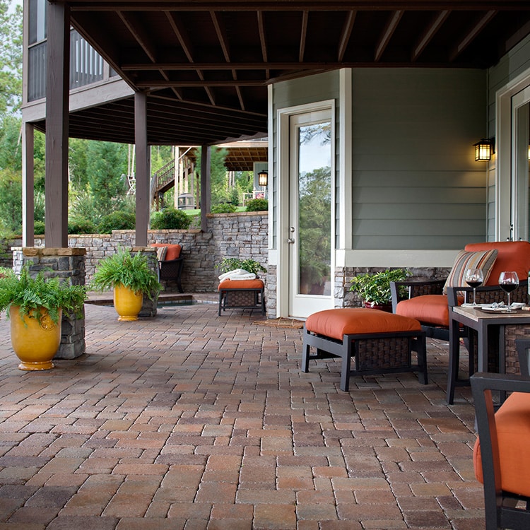 A brick patio with orange cushioned chairs and potted plants, adjacent to a house with gray siding and large windows, under a covered balcony.