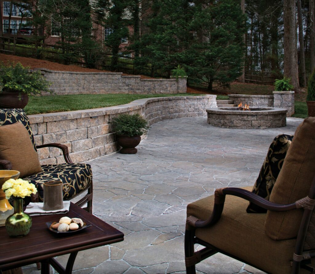 A stone patio with cushioned chairs, a small table with snacks and flowers, a curved retaining wall, potted plants, and a fire pit surrounded by trees.