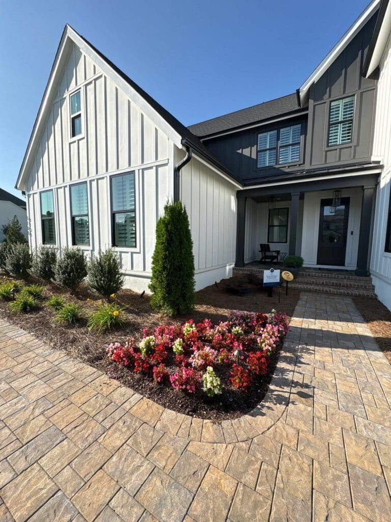 Single-story modern farmhouse with white and dark gray siding, a brick walkway, landscaped garden beds, and a small front porch with seating.