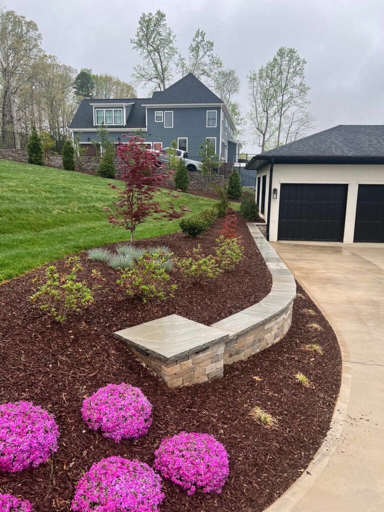 A landscaped front yard with pink flowers, a curved stone bench, mulched garden beds, and a gray house with a detached black garage in the background.