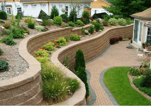 A landscaped backyard with curved tiered retaining walls, decorative plants, and a brick patio pathway beside a house under construction.