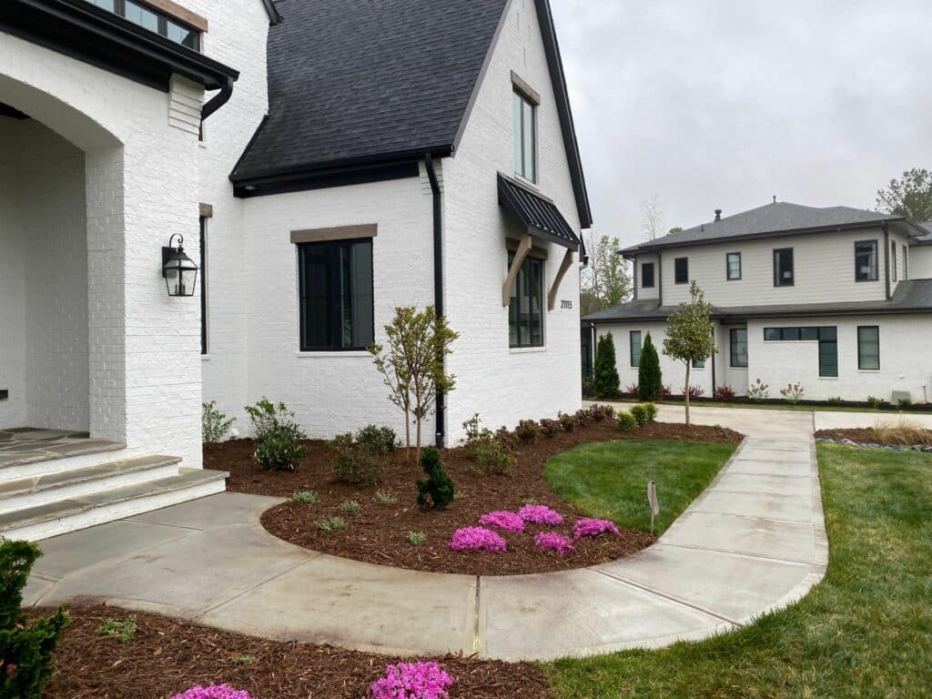 A concrete sidewalk curves through a landscaped yard with pink flowers in front of two modern white houses on a cloudy day.