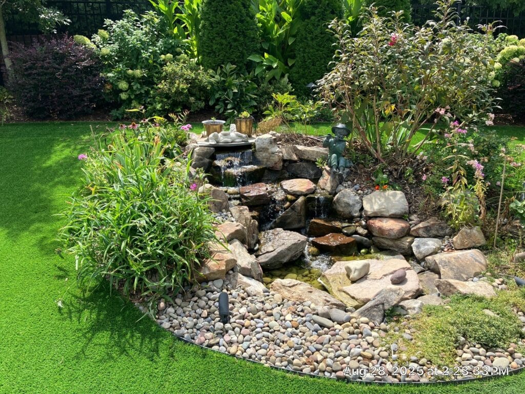 Small backyard garden with decorative rocks, a water feature, various green plants, and artificial turf under bright sunlight.