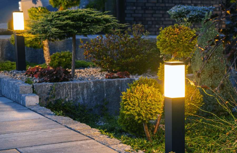 Two illuminated outdoor garden lights line a stone pathway next to neatly trimmed bushes and small trees.