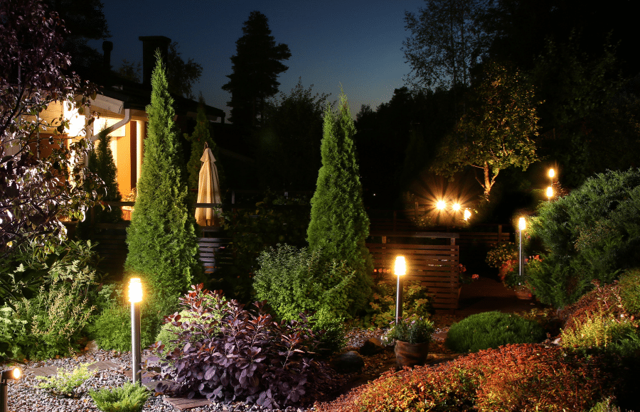 A landscaped garden at dusk with illuminated pathway lights, various plants and trees, and a lit-up house in the background.