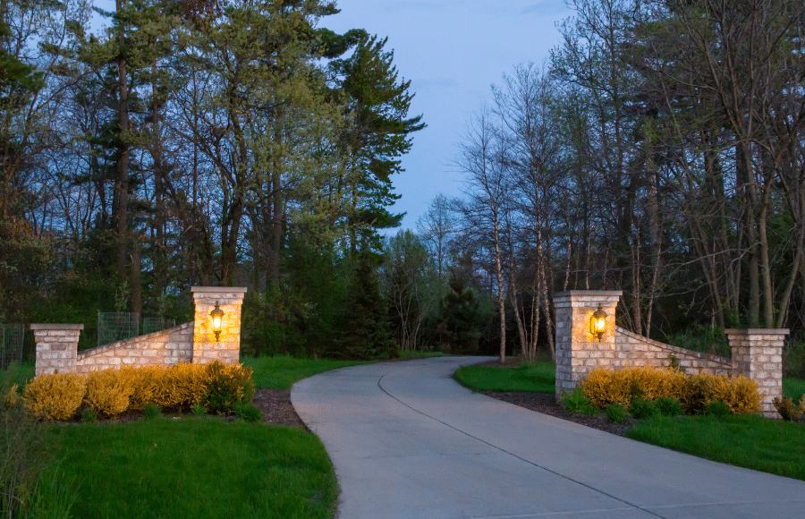 A paved driveway curves between two stone pillars with lanterns, surrounded by green grass, yellow shrubs, and trees at dusk.