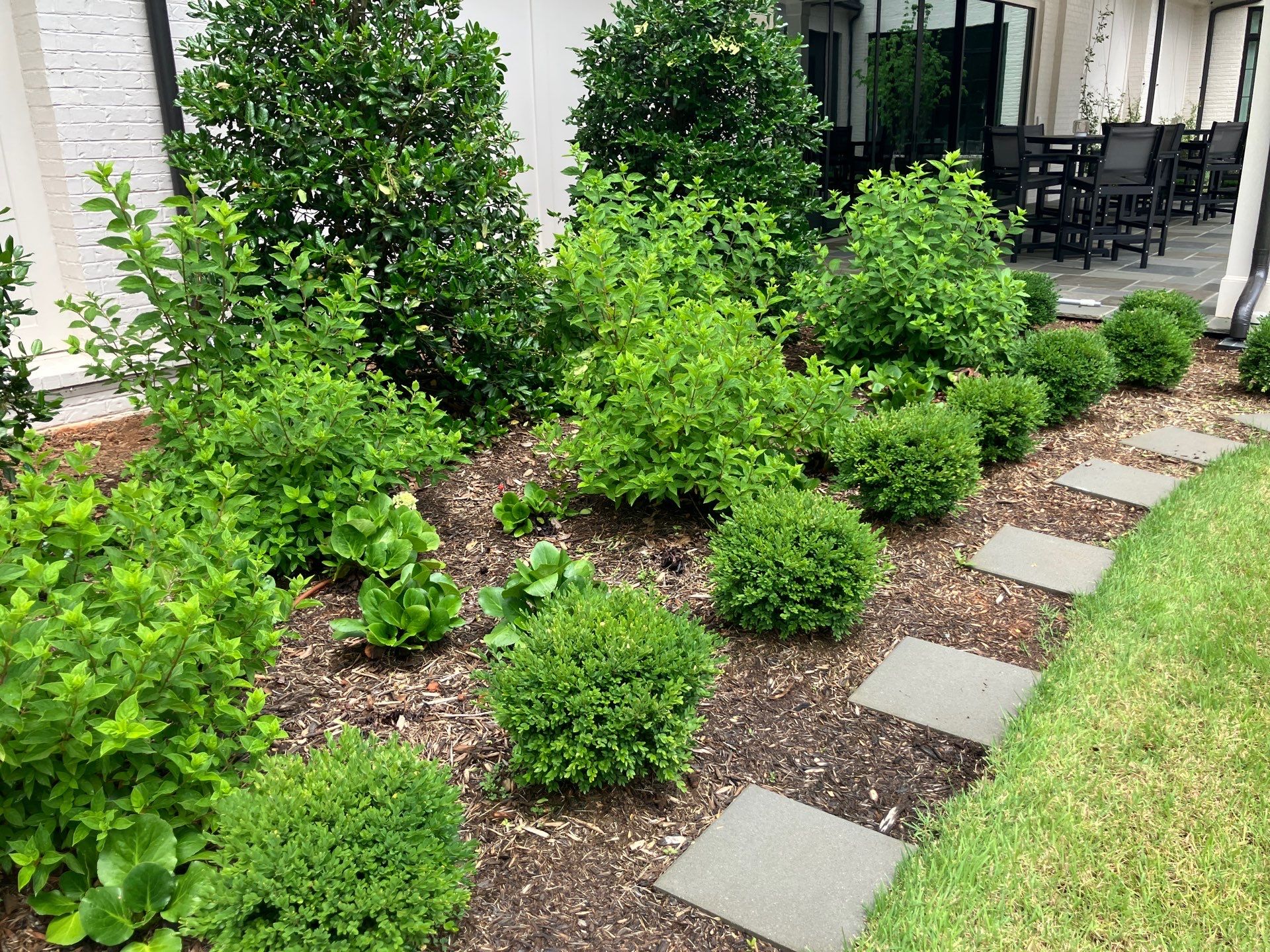 Green shrubs and bushes are planted in a mulched garden bed beside a patio with black outdoor furniture and large windows; stone pavers form a path along the edge.