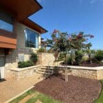A landscaped yard with stone retaining walls, mulched garden beds, green grass, a flowering tree, and a modern house with large windows in the background under a clear sky.