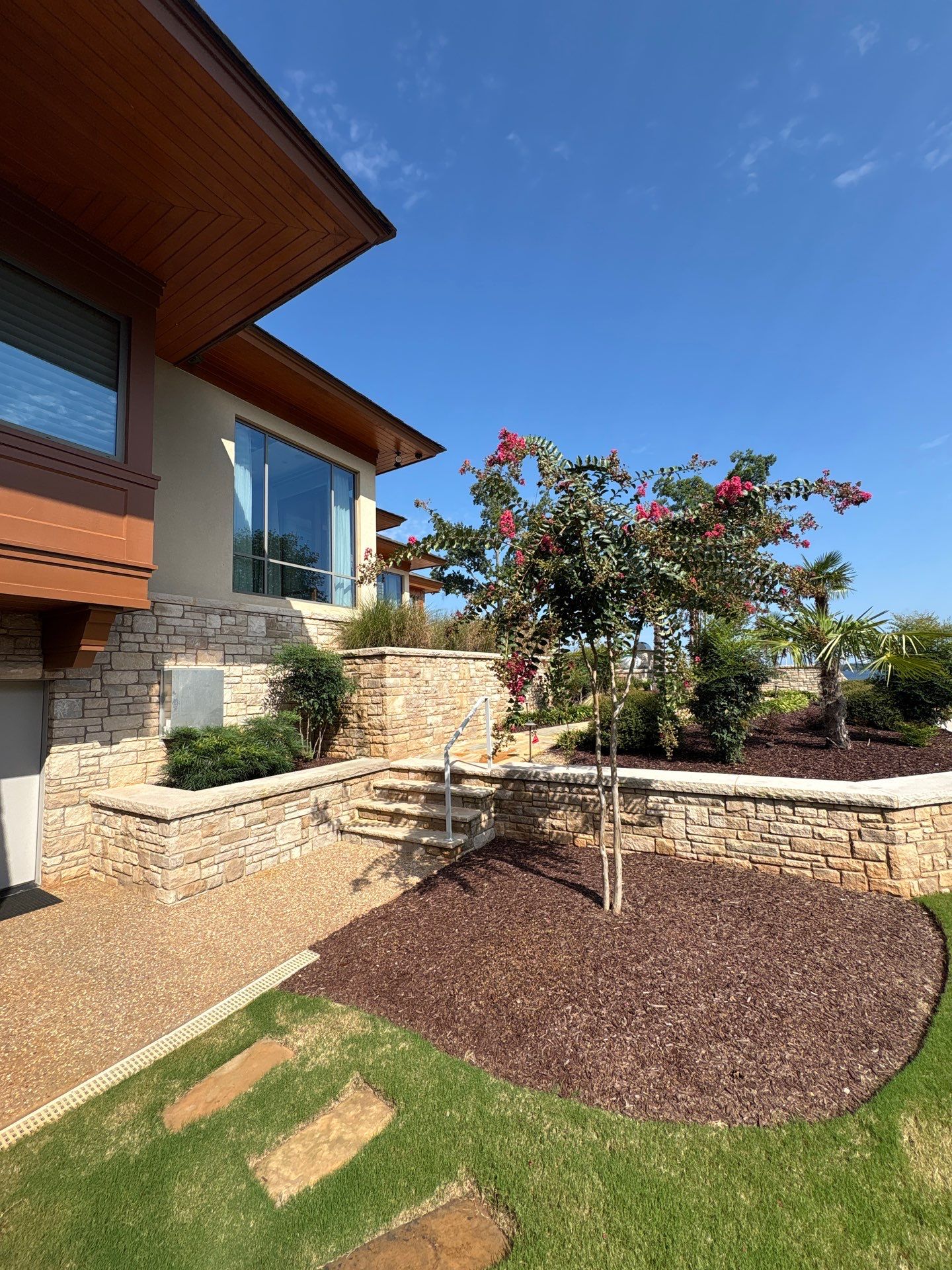 A landscaped yard with stone retaining walls, mulched garden beds, green grass, a flowering tree, and a modern house with large windows in the background under a clear sky.
