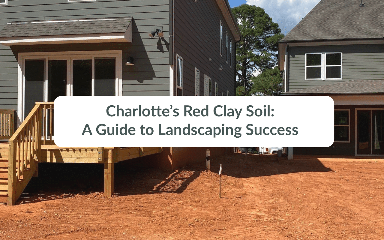 Two modern homes with gray siding sit on lots of exposed red clay soil under a blue sky, with a deck extending from one house.