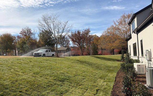 A white pickup truck waters a grassy lawn with a hose near a beige house, surrounded by trees with autumn foliage under a partly cloudy sky.