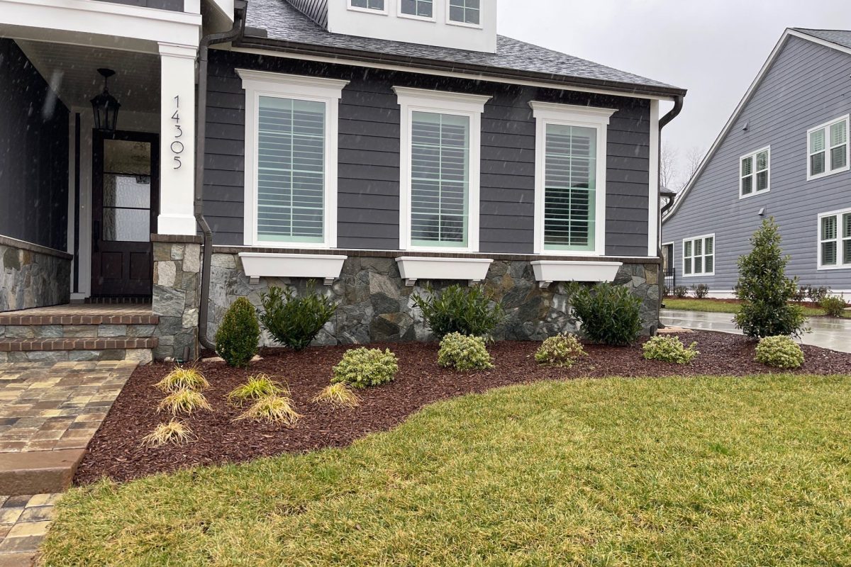 A modern gray house with white trim, three front windows, stone foundation, landscaped shrubs, and a neatly edged lawn on a cloudy day.